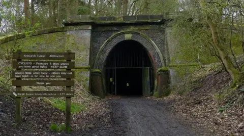 MAT FASCIONE/GEOGRAPH Entrance to tunnel at which iron gates are just visible. There is a grey stone archway over the entrance. There is a steep grass slope to the left and a steep mud slope to the right. A sign to the left has instructions for walkers and cyclists.