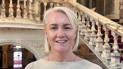 Tracy Kelly standing in Belfast City Hall, she is wearing a cream top and has blonde hair