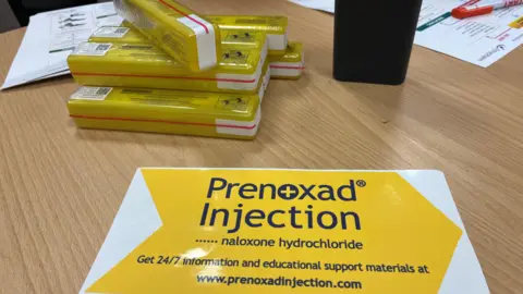 A pile of naloxone kits on a table next to a yellow and white promotional mouse mat showing where to get information.