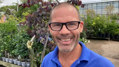 A man with very short brown hair and brown and grey facial hair is smiling. He is wearing a blue t-shirt and there are plants behind him on shelves in an outdoor area