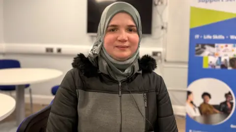 BBC Sabah, she is wearing a grey headscarf and a black and grey coat and is sitting in a room with white walls. Behind her a white table and blue chair can be seen.