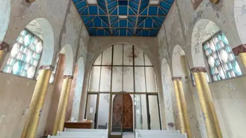 Image shows the interior of a church, with gold columns along the walls and stained glass windows high up on the walls. There are pews down the length of the church leading to the altar. 