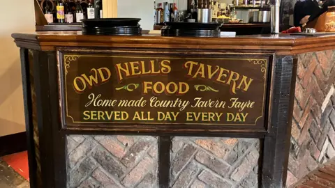 A sign for Owd Nells Tavern saying "Food homemade country tavern fayre served all day every day". It's affixed to the end of the bar, bottles can be seen towards the top of the image.