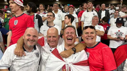 Paul Wilson Jersey football fans Paul Starkey, Dave Houze, Paul Wilson, and Tony Rogers at the Azteca Stadium in Mexico after England's World Cup win. They are smiling and are wearing red and white England shirts. Dave Houze has a red and white England flag round his shoulders. Other England fans can be seen in the stadium behind them.