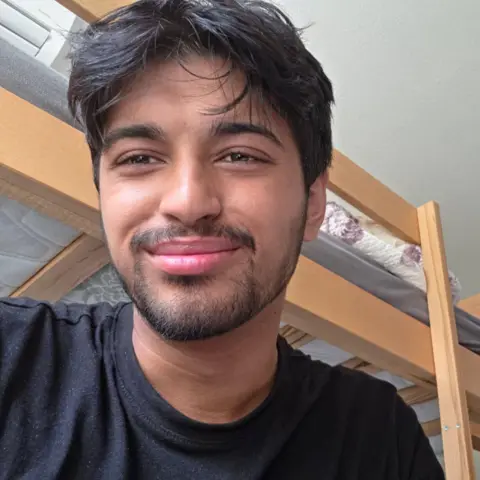 Haadi Nasir A man with black hair and a black beard smiling, he is wearing a black top and behind him is wooden bunk beds. 