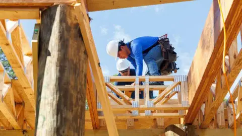 Getty Images Workers wearing hard hats stand on a wooden house frame, measuring and securing beams as part of a construction build.