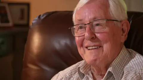 BBC Stephen Harrington is an older man, aged 83. He has white hair and wears thin-rimmed clear-lensed glasses. He is sitting on a brown leather chair and wears a white and brown checked shirt. He is smiling.