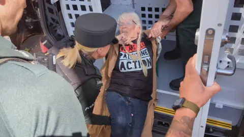BBC A woman with white hair being moved into the back of a police van. Two police officers are next to her. 