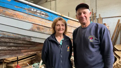 Julie and Ian Coultas are standing by the coble Imperialist in the workshop where she is being restored.