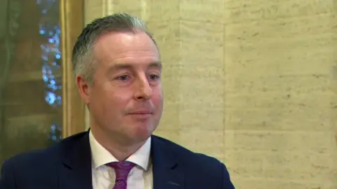 Givan is wearing a navy suit with a white shirt and a purple tie. He's standing in the lobby at Stormont parliament building. There are Christmas lights behind him. 