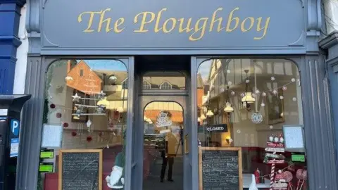 The front of a cafe, which has two large windows and the frames and door are painted grey. There are Christmas decorations in the windows