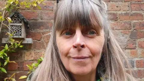 A woman with long brown hair stood in front of a brick wall with a plant growing up it