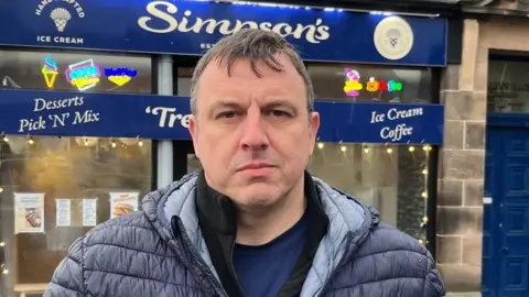 A man with short grey hair wearing a parka jacket standing in front of a shop with blue signage