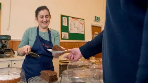 Ebenezer Church A woman behind a cake counter at Ebenezer Church in Horfield in Bristol serves a man with a slice of cake. She is smiling and wearing a blue apron