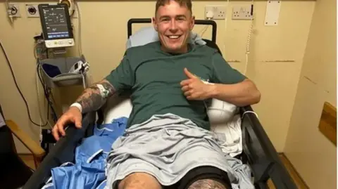 Jack Jarvis Jack Jarvis in a hospital bed attached to machines with something wrapped around his leg. He has a thumb up and is smiling.
