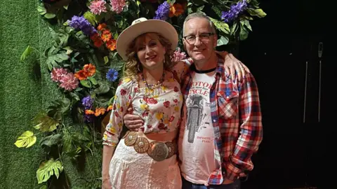 Maxine and Bryan Day A woman in a hat and floral blouse, with arms around a man in a checked red and blue shirt and motorcycle t-shirt