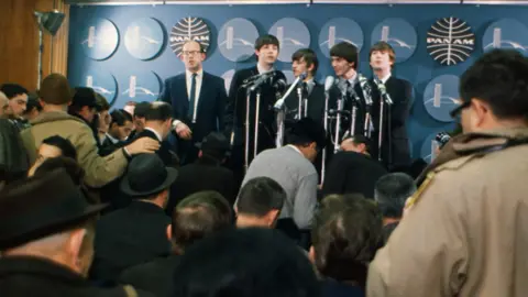 Getty Images The Beatles stood behind microphones doing a press conference in front of a room full of journalists.