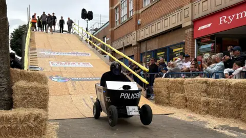 Barrow BID A person drives a soapbox derby car that has been painted to look like a police car down a large wooden ramp. At either side of the ramp are straw bales and people stand at the top of the ramp watching the racer. Crowds line the streets watching the kart go past.