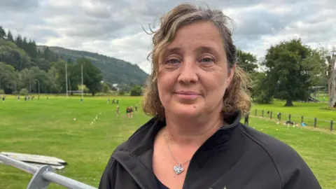 Elaine Jones with the rugby pitch in the background. She has brown curly hair half tied back and is wearing a black jacket and a silver necklace pendant in the shape of a heart.