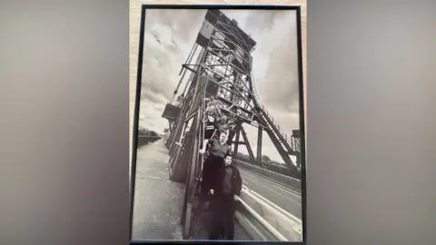 Bridgehill Day Centre A black and white picture of a photograph of a line of men descending the steps of a tall metal bridge. The structure is above a road and has a pavement on either side. The men are looking solemnly into the distance and the sky is cloudy overhead.