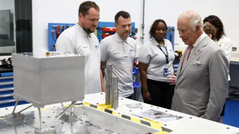 PA Media King Charles III during a visit to Harwell Science and Innovation Campus in Didcot, he is wearing a grey suit and is surrounded by people wearing the same white t-shirt uniform. Charles is looking at some scientific equipment.