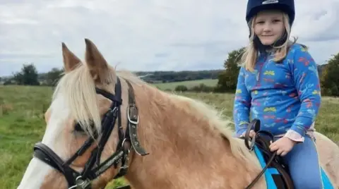Jackson family A young girl sits on a horse and smiles at the camera. She has long blonde hair and wears a riding hat, blue leggings and a bright blue top with colourful patterns. 