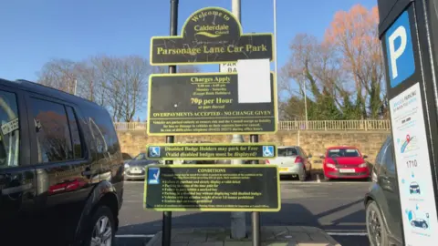 Nathan Turvey/BBC A picture of a car park sign with prices on it in Brighouse, with cars parked either side and behind.