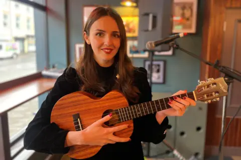 Amelia Coburn, who has long brown hair and red lipstick and his wearing a black jumper, is holding a small wooden acoustic guitar and is smiling.