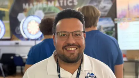 Elliot Deady/BBC A man with glasses and a landyard around his neck 