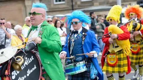 Pacemaker Band members holding colourful drums and bagpipes wear blue and yellow wigs as they take part in Belfast Pride Parade. The person in the front is wearing a green blazer, a green tartan skirt and green sunglasses. In the middle, someone else is wearing a blue wig, a blue blazer, vest and tartan skirt and in the back are two people wearing yellow wigs, a red blazer and a yellow tartan skirt.