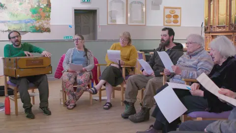 Hopscotch A man in a beard and glasses leads five other people - four women and a man - in song. They look like there is an organ behind them and they are all holding sheets of paper apart from the man leading them who appears to be playing some kind of musical instrument, a wooden box on his knees.