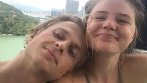 A couple look at the camera for a selfie. They appear happy and smiling. The man has blond hair and is leaning on the shoulder of the woman who has fair hair which is tied back from her face.