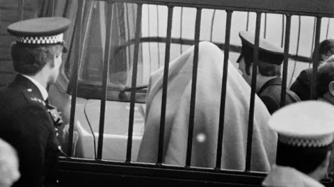 A black and white image of Malcolm Fairley captured on 27 February 1985. He has a blanket over the top half of his body and his back is facing the camera. He is flanked by several police officers and is in a gated compound at St Albans Crown Court. In front of the gate, there is an officer wearing a white hat.