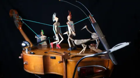 Robert Race A violin with small models of humans and birds on top of it. They are attached to the violin with string, allowing them to be manouvered.