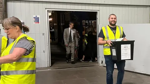 SIMON DEDMAN/BBC A man walking in carrying a big black box which is a ballot box into a building that looks like a warehouse. He is smiling and wearing a high-viz jacket.