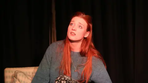 A young red-headed woman in a grey jumper sits on a chair on a black stage, looking into the distance.