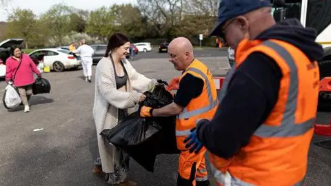 Residents hand rubbish to refuse workers at a Mobile Household Waste Centre in Senneleys Park on 8 April. One woman in a long beige cardigan is handing over two black bin bags and another in a pink coat is walking up with bags to deliver.