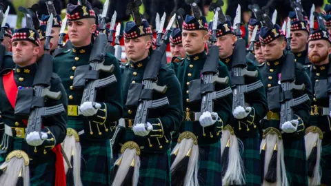 Members of the Royal Regiment of Scotland during a military procession. They are wearing kilts while marching with guns