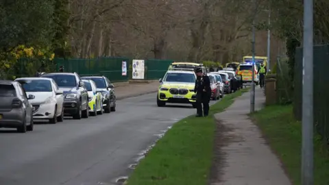 Qays Najm/BBC Cars parked on either side of the road on Laundry Lane. There are marked police cars in attendance and police officers walking about.