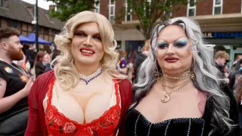 A drag queen with blonde hair and a red dress, next to a drag queen with grey hair and a black dress in a large crowd of people