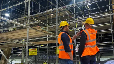 Emma Baugh/BBC Two workers wearing yellow hard hats and orange high-vis jackets are standing next to each other, having a conversation. One of them is pointing upwards towards some scaffolding. 