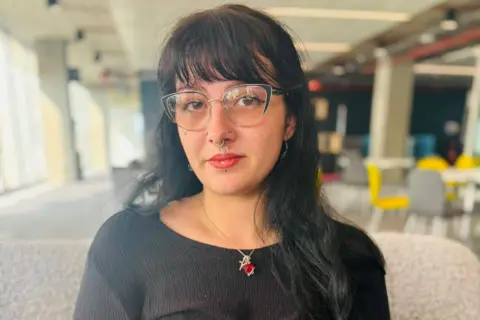 Meaghann Murphy, a woman looking at the camera, she has long dark hair, and is wearing glasses and a black top, and a necklace, pictured in a large room setting.