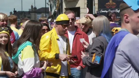A group of people wearing fancy dress. One man dressed as Ali G is being asked for identification