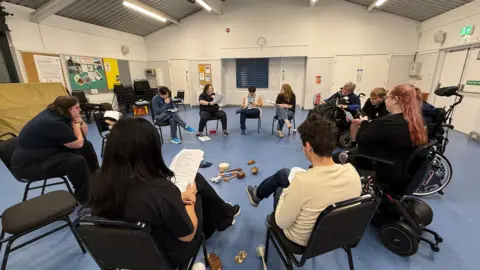 Steve Hubbard/BBC People sitting in a circle in a village-hall style room.
