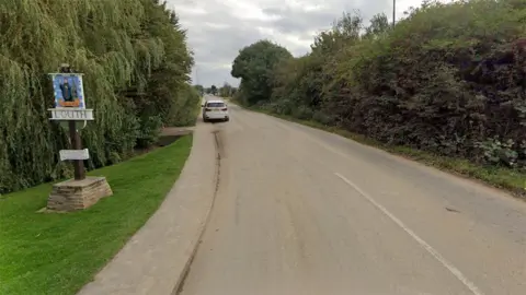 Brackenborough Road as it enters Louth a small road with hedges either side and a white car parked on one kerb. In the front is an ornate sign for Louth mounted on a wooden pole and stone base 