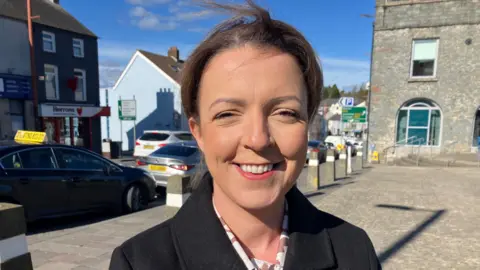 Infrastructure Minister Liz Kimmins smiling at the camera.  She has dark hair tied back and is wearing a black coat over a white and pink patterned blouse.