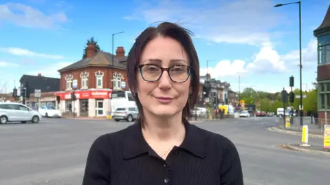 A woman with a black shirt and glasses looking into the camera. She is standing in front of a junction. She has a little smirk but looks quite serious. 