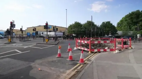 The junction of Princes Avenue and Spring Bank West in Hull. The road, which features multiple sets of traffic lights, is covered with orange and silver cones. A red and white barrier surrounds a large hole and a large pile of rubble. An industrial building stands in the distance.