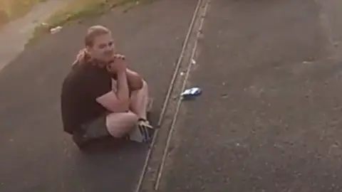 A man with hair in a pony tail sat cross-legged on the pavement.