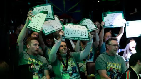 MEWC crowds of people holding signs with Excel-related messages showing support for their favorite contestant. one reads "excel wizard"and the other one says "Jack Excels."They are all covered in green light, which spreads across the crowd and onto the field.